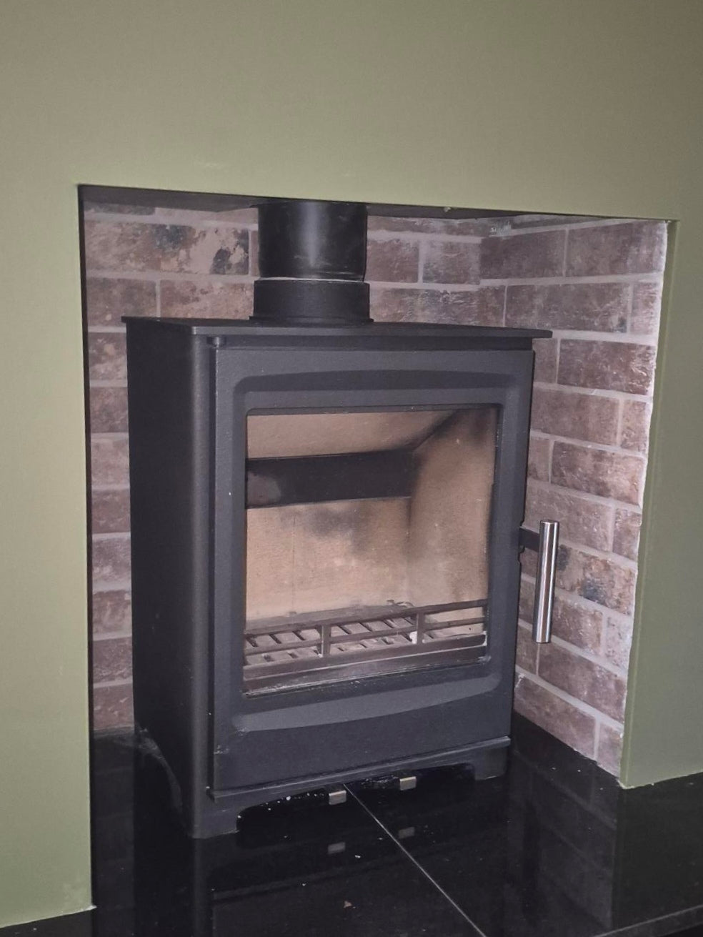 Black wood stove installed in a brick wall with a green wall background Lucan Stove San Remo Wood Burner Multi Fuel Solid Fuel & Chimney Lining Kit Bundle 5kw