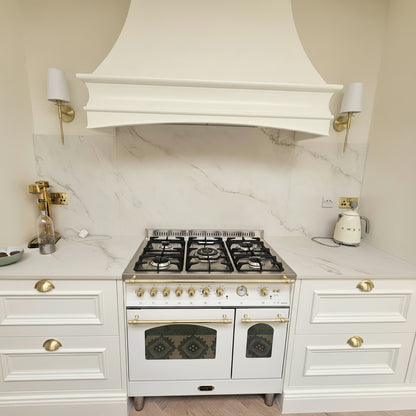 A white gas range stove with gold handles and knobs, set under a white range hood in a kitchen with white marble backsplash and white cabinets with gold hardware- with a Lofra 90cm White and Brass Finish