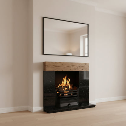 A modern gas fireplace insert with a black marble surround and a light wood mantel, shown lit with orange flames. A large black-framed mirror hangs on the wall above it in a clean, minimalist room with beige walls and light wood flooring.