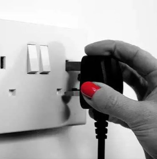 A close-up of a hand with bright red nail polish plugging a black electrical adapter into a white wall switch outlet.