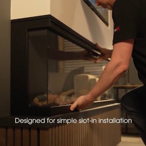 A man in a black t-shirt is installing a modern black wall-mounted fireplace insert into a wooden base. The image includes the text 'Designed for simple slot-in installation' at the bottom. Atmos Fire FX Versa Range 