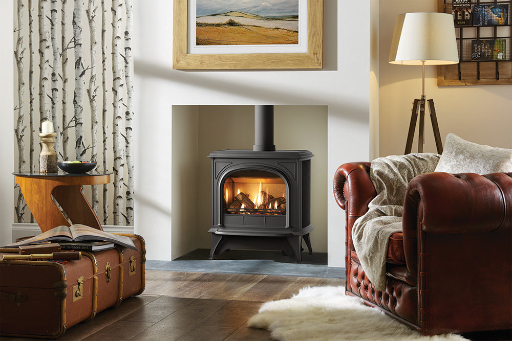 A conventional Gazco Huntingdon 40 clear glass gas stove with a glass front and a log flame effect, installed in a fireplace alcove with a framed picture and mirror above, in a room with a leather sofa and a fur throw.