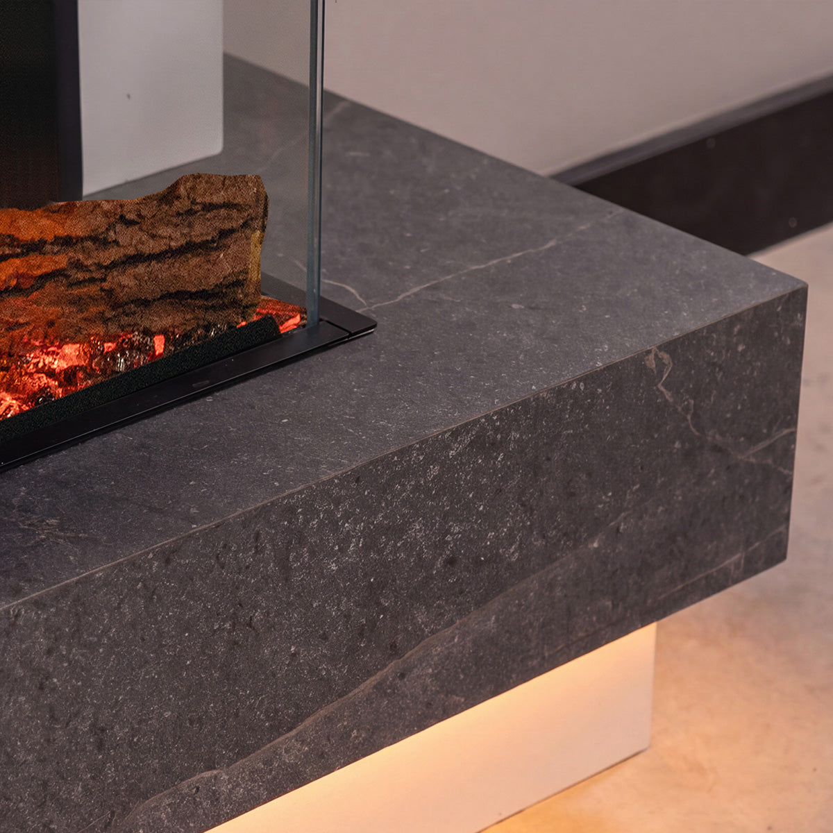 A modern floating fireplace mantel made of dark gray stone, featuring a glass wind guard and a faux log with glowing embers inside.