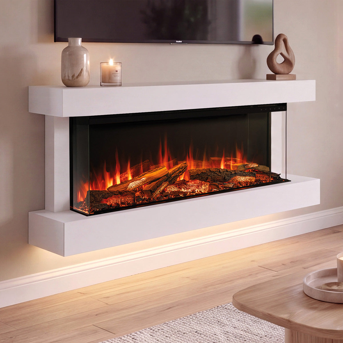 A modern wall-mounted electric Turin 120 fireplace with a white floating mantel and glowing faux logs, shown installed in a living room setting with a TV above, candles on the shelf, and a wooden floor below.