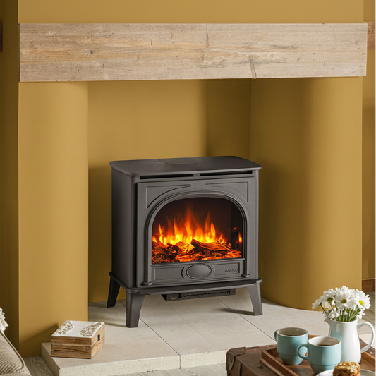 A black wood-burning Electric Stockton2 Medium Stove with a glowing orange and yellow fire inside, set in a rustic living room with mustard yellow walls, a stone hearth, and a brown leather trunk coffee table.