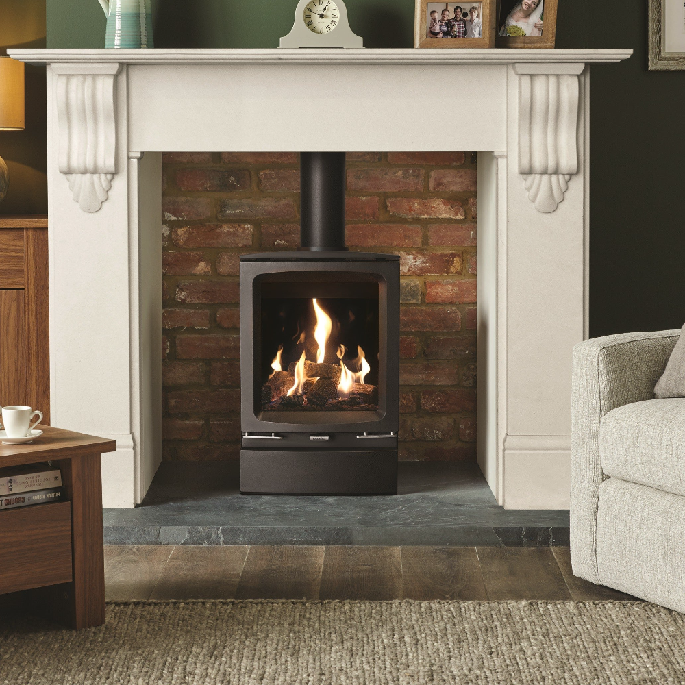 A black Gazco Vogue Midi gas stove with a realistic log effect fuel bed, installed in a contemporary style interior with a decorative brick wall and a polished stainless steel insert, alongside a programmable thermostat.