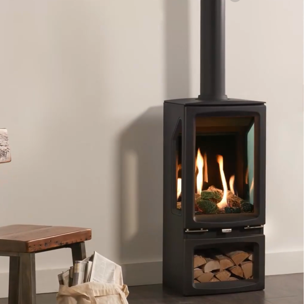 Modern Gazco Vogue Midi T black freestanding stove with LOG BOX fire burning inside, next to a wooden chair and a small table.